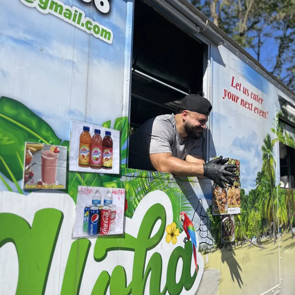 A crew member leaning out the Aloha truck's service window handing over an order, the catering phone number painted beside him