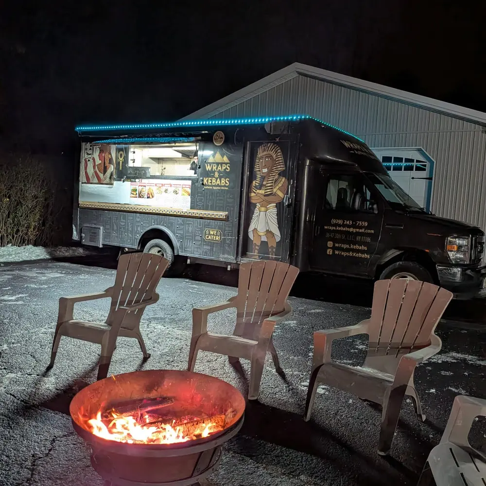 The Wraps & Kebabs truck lit up at night behind three empty Adirondack chairs around a fire pit