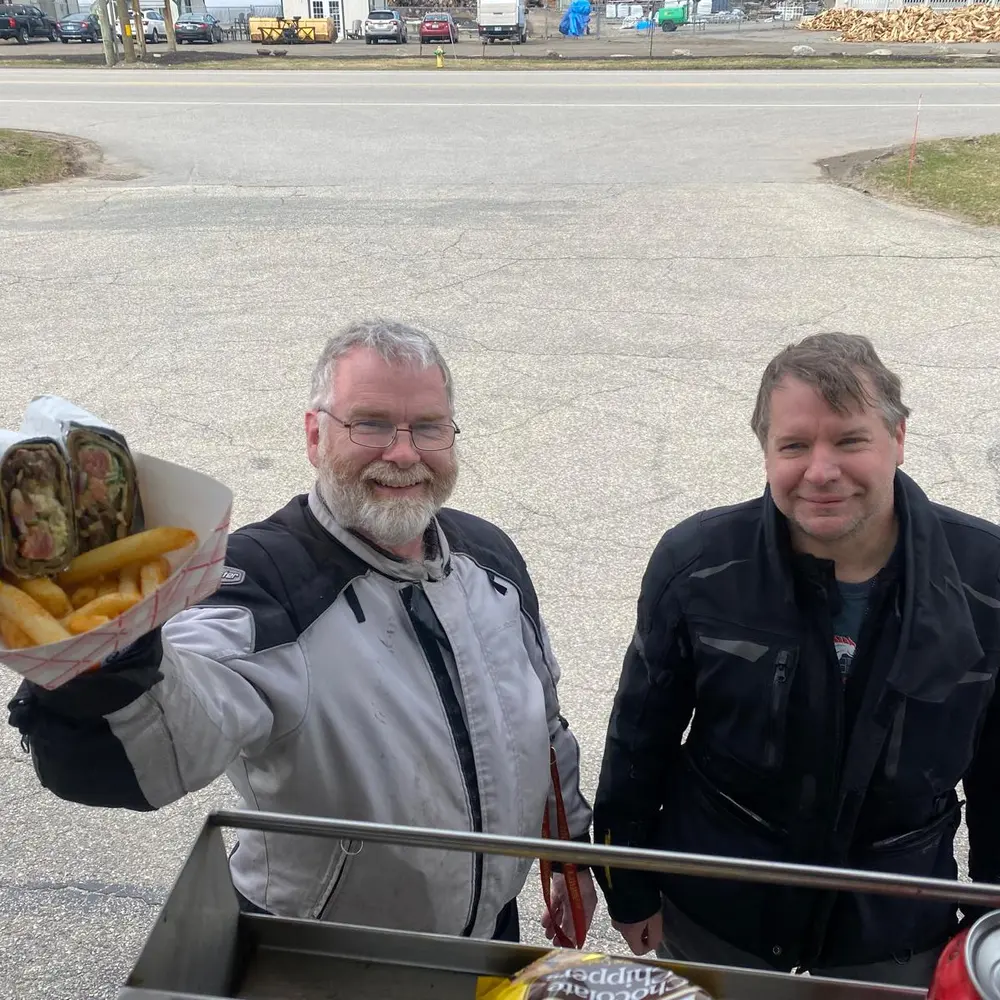 Two motorcycle riders smiling and holding a takeout container of food at the truck