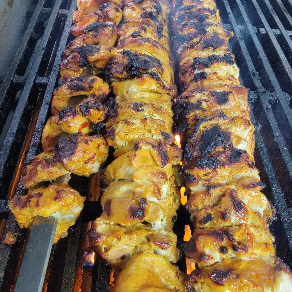 Three steel skewers of marinated chicken kebab charring over the grill