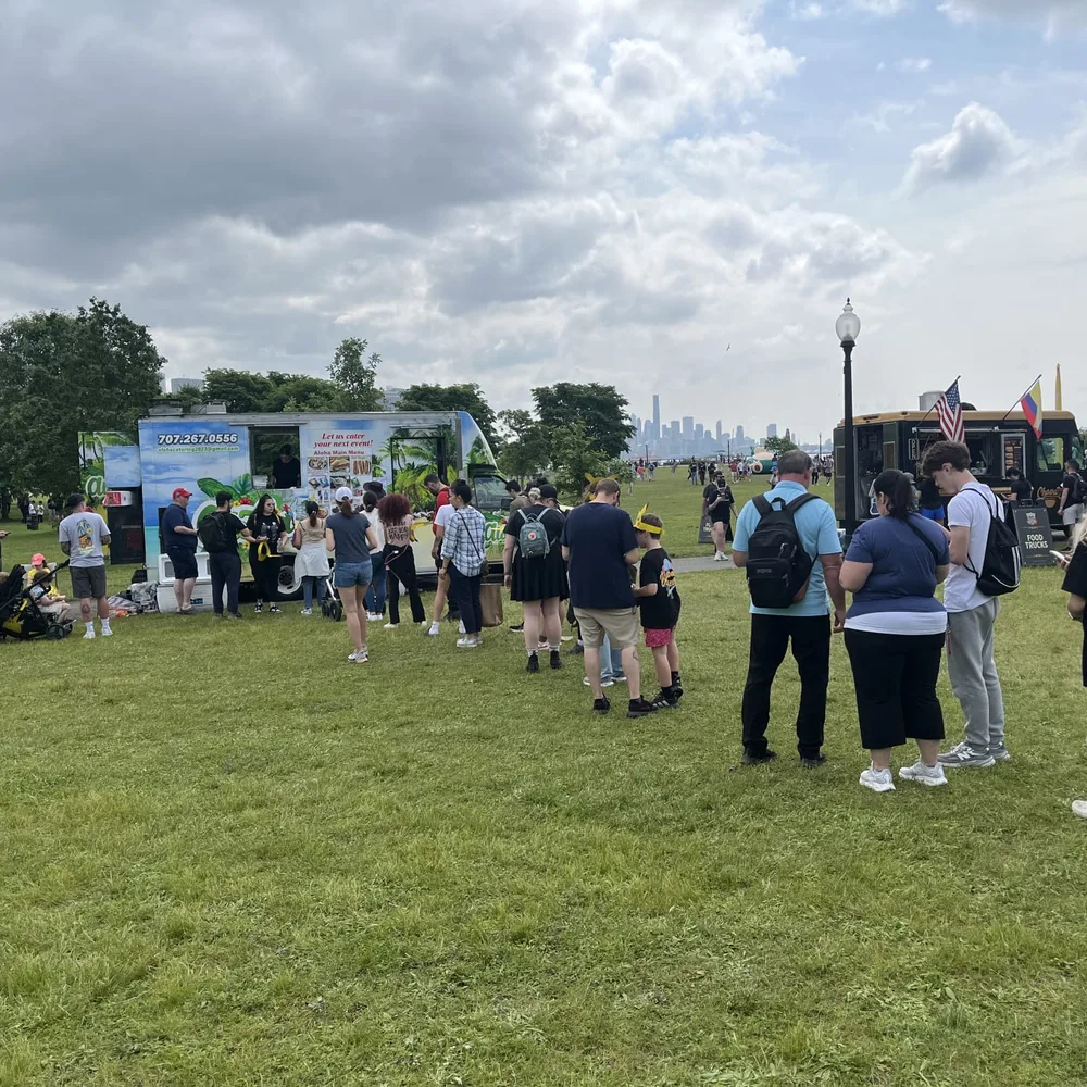 A long line across the grass to the Aloha truck at a waterfront festival, the Manhattan skyline behind