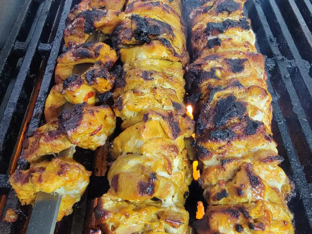 Three steel skewers of marinated chicken kebab charring over the grill