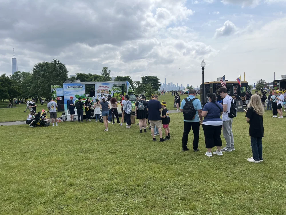 A long line across the grass to the Aloha truck at a waterfront festival, the Manhattan skyline behind